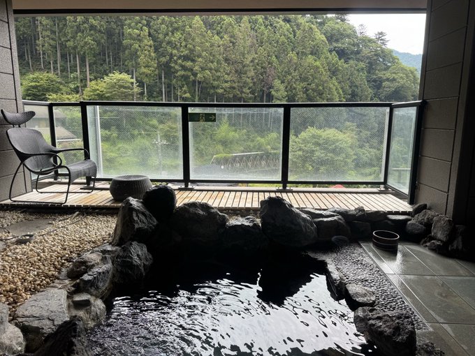 An indoor hot spring or onsen bath surrounded by rocks, featuring a wooden deck and chair, overlooking a green forested mountainside through large windows.