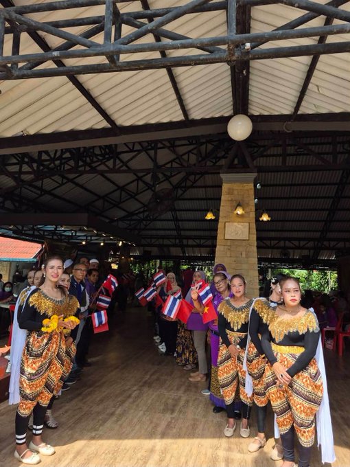 Dancers in traditional Thai attire hold small Thai flags at what appears to be the opening celebration of the Pi-GCV Peace Alliance's 2nd branch in Krabi, Southern Thailand. The festive gathering takes place under a large open-air pavilion with a wooden truss roof, honoring the team's milestone achievement.