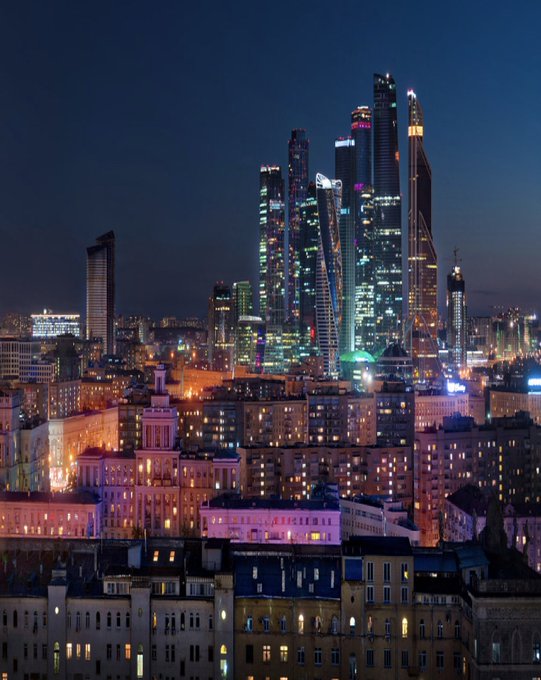 A nighttime aerial view of Moscow's illuminated skyline showcases the Moscow City financial district skyscrapers towering over older residential and Stalinist-era buildings below. The image visually anchors the news of MOEX's Moscow Exchange launching retail cryptocurrency spot trading, with the modern financial center in the background symbolizing Russia's expanding crypto infrastructure.