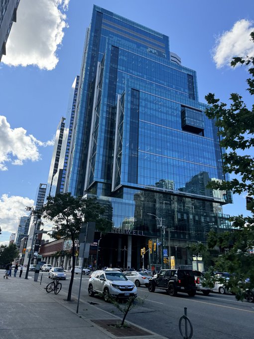 A modern blue-glass skyscraper towers over a tree-lined city street, with cars passing by on the road below. The image reflects a relaxed Sunday urban scene, complementing the "happy Sunday" message by capturing the calm, sunny atmosphere of a city weekend with its bright skies and gentle street activity.