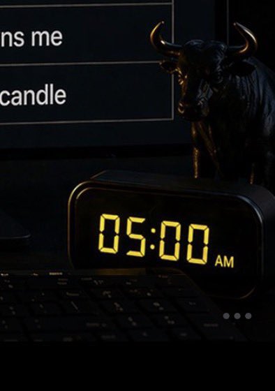 A digital clock displays "05:00 AM" in yellow LED digits on a dimly lit desk beside a keyboard and a bull figurine, symbolizing an early-morning bullish alert. The "ANS ME" and "candle" text in the background ties to the post's mention of a potential $ANSEM listing on Binance and bullish price expectations.