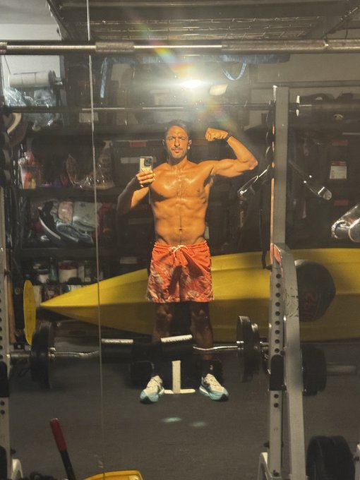 A muscular man takes a mirror selfie in a gym at 5AM, flexing his bicep while wearing orange shorts and sneakers, capturing a pre-event pump before heading to RareEvo to cover blockchain and AI topics.