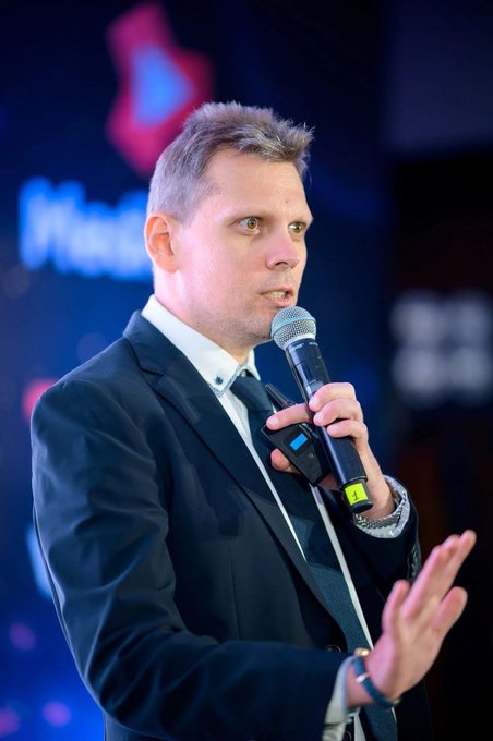 A man in a dark suit and tie holds a microphone while speaking, with a partially visible "Nas..." logo in the background. The image relates to the post about Sergey Sevantsyan joining UXUY as CMO, likely showing him at a speaking engagement or conference.