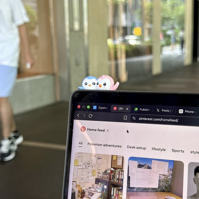 A laptop displaying the Pinterest homepage, with two small cartoon penguin figurines (blue and pink) perched on top of the screen.