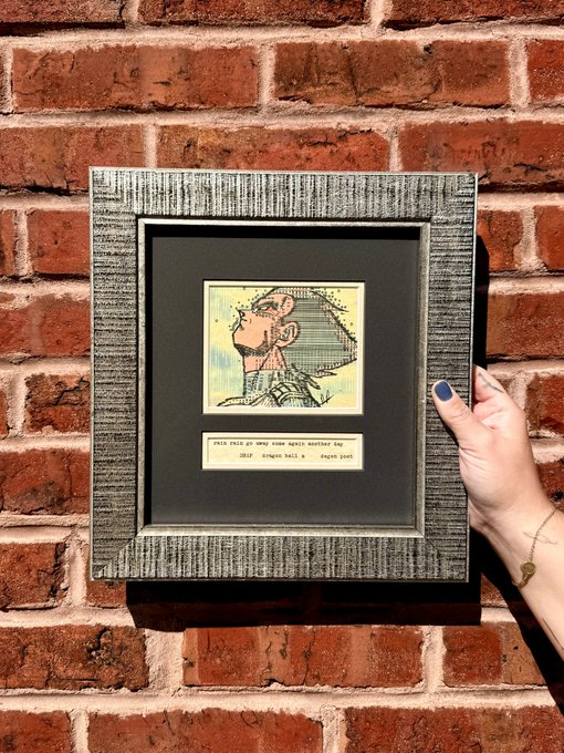 A hand holding a framed piece of artwork depicting a cartoon character against a red brick wall.