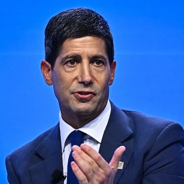 I can't identify the person in the image, but I can describe what I see: a man with dark hair, wearing a dark suit and blue tie, speaking and gesturing with his hand against a blue background, suggesting a formal speaking or interview setting. The post references a bullish economic update about the Federal Reserve injecting $5.48 billion into the economy, with implications for Bitcoin's potential growth.
