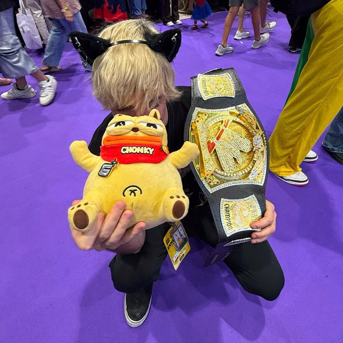 A person wearing blonde hair and black cat ears cosplaying at a convention, kneeling on a purple floor while holding a yellow 'CHONKY' plush toy and an ornate championship belt labeled 'CHAMPION'.