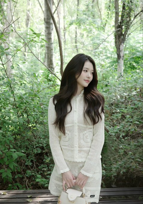 A young woman with long dark wavy hair in a white crochet top and skirt, holding a small white handbag while standing in a lush forest setting.