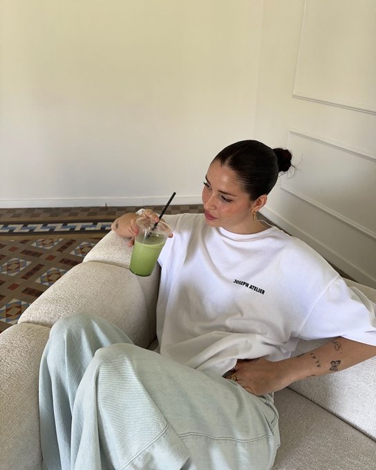 A woman with dark hair in a bun sits on a beige couch holding a green iced matcha drink with a straw, wearing a white t-shirt that reads 'JOSEPH ATELIER' and light gray sweatpants.