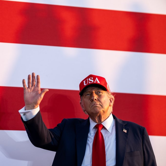 Donald Trump, wearing a red "USA" cap and dark suit with a red tie, waves in front of an American flag backdrop as he indicates openness to government Bitcoin acquisitions.