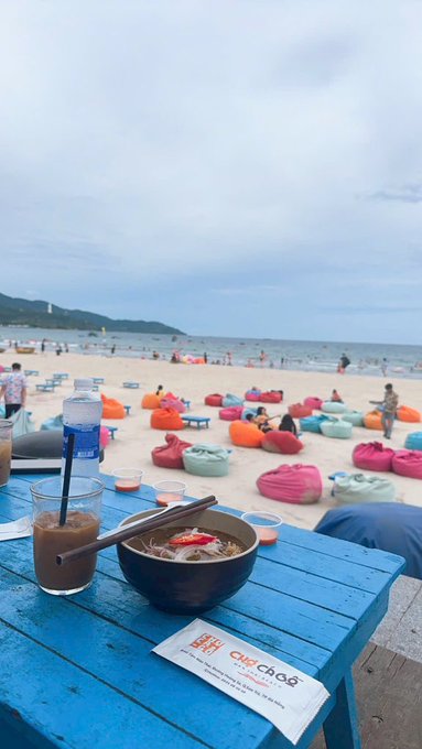 A first-person view of a meal on a blue wooden table at a beach, featuring a bowl of noodles, an iced coffee drink, a water bottle, and chopsticks, overlooking a sandy beach filled with colorful bean bags.