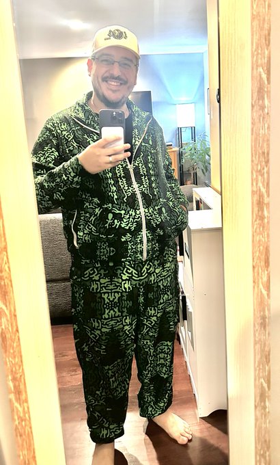 A smiling man with a beard, glasses, and a beige cap takes a mirror selfie wearing a green patterned onesie in a home interior.