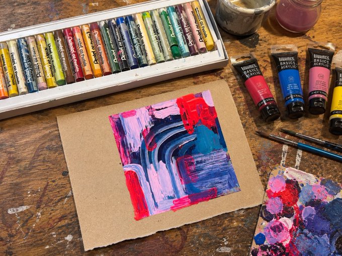 An artist's workspace with a colorful abstract painting in progress on brown paper, surrounded by boxes of Caran d'Ache Neocolor II crayons and Liquitex Basics acrylic paint tubes.
