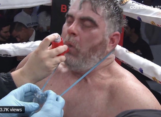 A close-up shot in a boxing ring corner where a trainer wearing blue gloves sprays a substance from a red aerosol can into the mouth of a sweaty, bearded boxer, with '3.7K views' and a Coindesk ad visible.