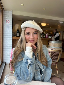 A young woman wearing a white beret and denim jacket smiles at the camera in what appears to be a French bistro, with wine bottles and a wooden barrel visible in the background. The beret and bistro setting tie perfectly to her post about exploring France and supporting "Les Bleus," while the mention of BC.Game aligns with the casual café atmosphere as she reminisces about her trip to Cannes.