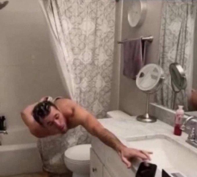A man is shown leaning out of a bathtub, scrubbing his hair with one hand while reaching for a phone on the bathroom counter, with a mirror and shower curtain visible.