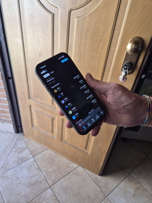 A hand holds a smartphone displaying a crypto portfolio app in front of a wooden door with a brass doorknob, showing various token balances and 'Top tokens' list.