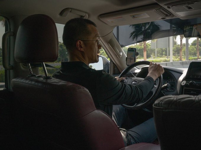 A man wearing glasses is driving a car with leather seats, glancing at a smartphone displaying a navigation map mounted on the dashboard. The image illustrates the post about earning many new driver's licenses, showing the focus and concentration required behind the wheel.