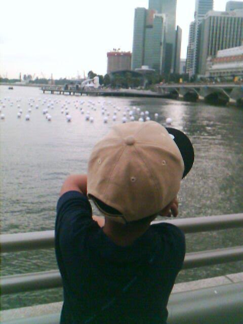 A child wearing a tan cap stands with their back to the camera, looking out over a body of water dotted with white floats, with a city skyline in the background.