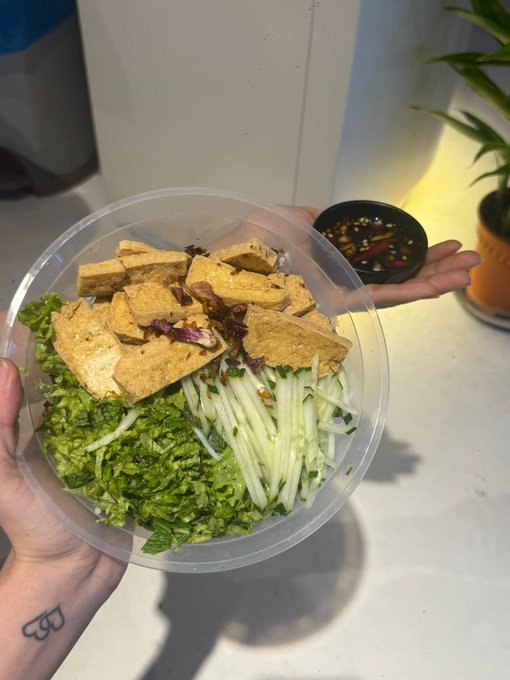 A person holds a plastic bowl containing Vietnamese-style fried tofu, shredded green papaya, and herbs, with a small dish of fish sauce dipping on the side.