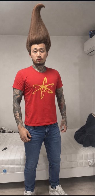 A heavily tattooed man wearing a red t-shirt with an atom symbol, blue jeans, and an extremely tall, voluminous brown hairdo, standing in a bedroom.