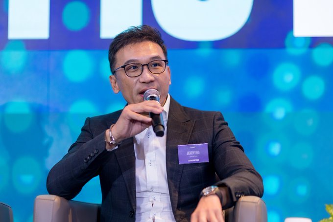A man in a dark suit and glasses speaks into a microphone while seated on a stage with a blue, bokeh-lit background, wearing a name tag.