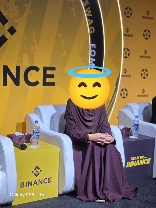 A person in a dark abaya seated on a white couch at a Binance event, with their face obscured by an angel emoji overlay; the background features Binance branding and a 'TEAM UP WITH BINANCE' podium.