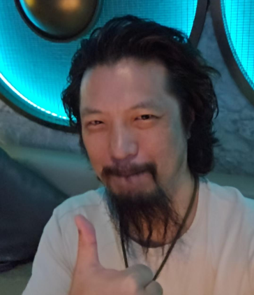 A man with a long dark goatee gives a thumbs-up in a blue-lit room.