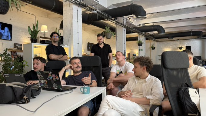 A group of people gather around a laptop in a renovated industrial-style coworking space in Belgrade, attentively watching as one person gestures with his thumb up. The scene captures the team's Friday coworking session, reflecting their close-knit community atmosphere as they engage in discussion, feedback, and collaborative growth.