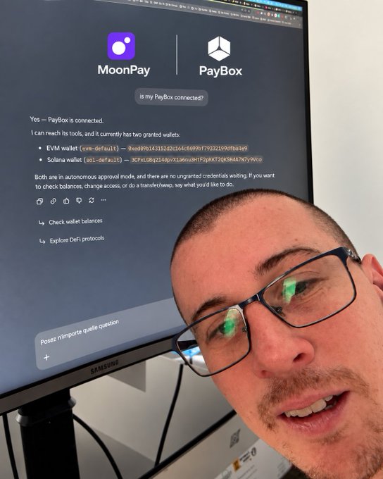 A man with glasses takes a selfie in front of a computer monitor displaying a chat interface with MoonPay and PayBox logos, showing a conversation about wallet connections including EVM and Solana wallets.