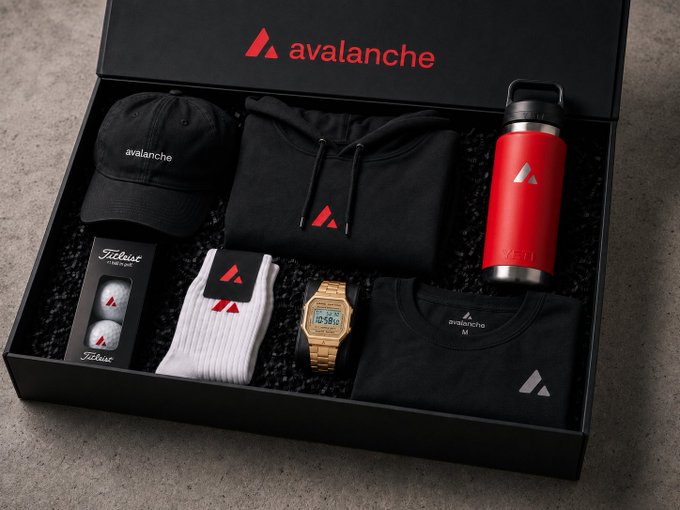 A black promotional gift box branded with the red "avalanche" logo contains a curated selection of merchandise including a black cap, hoodie, red YETI water bottle, Titleist golf balls, white socks, a gold watch, and a black t-shirt—swag items being offered as a giveaway ahead of the two-month countdown to Summit NYC.