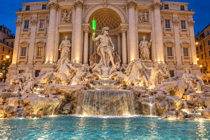 A large historic stone fountain with statues illuminated at dusk, water flowing from rocky elements into a pool.