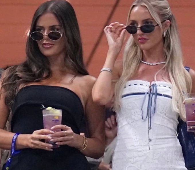 Two stylishly dressed women wear sunglasses at what appears to be an outdoor event, with one in a black dress holding a cocktail and the other in a white corset-style top adjusting her shades.