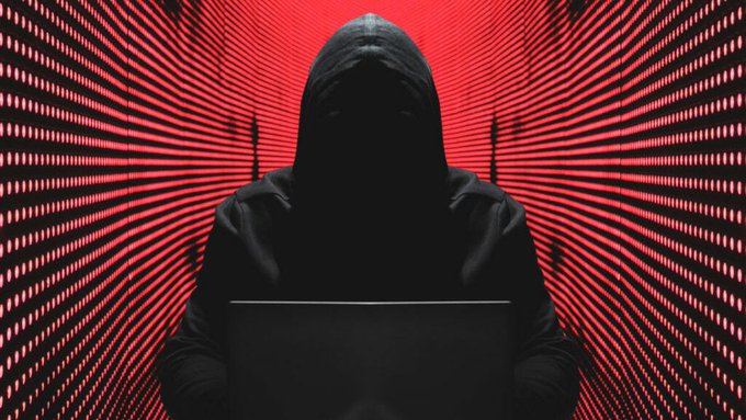 A stylized, dark image of a hooded figure obscured in shadows working on a laptop, set against a background of radiating red LED lines.