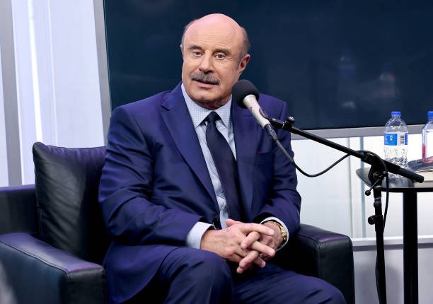 Dr. Phil McGraw, wearing a blue suit, sits in a black leather chair speaking into a microphone in what appears to be a studio setting.