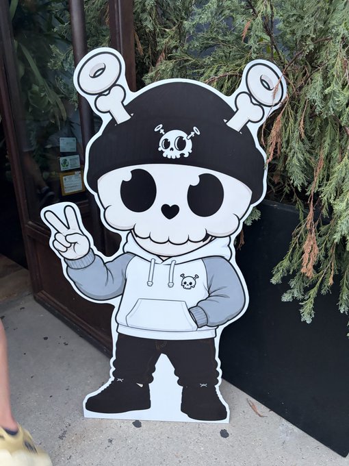An outdoor photograph of a large cartoon character cutout sign featuring a skeleton-like figure in a skull beanie with antennae, wearing a hoodie and making a peace sign, positioned near a building entrance with greenery in the background.
