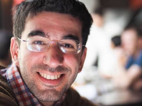 A close-up selfie of a smiling young man with dark hair, stubble, and rimless glasses, wearing a red and white checkered shirt.