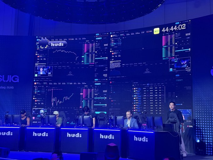 A large, curved screen displays cryptocurrency trading charts and data with a countdown timer at '44:44:02' during a 'hudi' (SUIG) event, with people working at computer stations below.