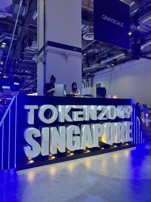 A registration desk at the TOKEN2049 SINGAPORE conference, featuring large illuminated lettering and a 'GRAYSCALE' banner hanging from the ceiling in a blue-lit venue.