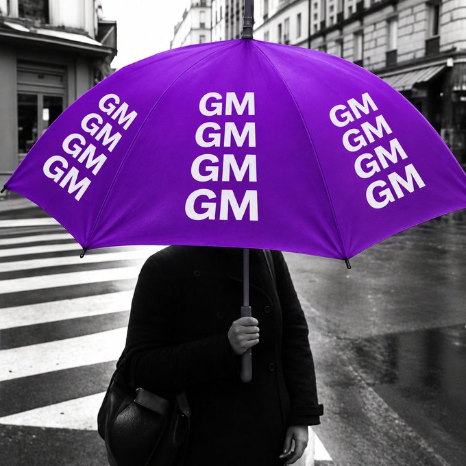 A black-and-white photograph of a person holding a bright purple umbrella covered in 'GM GM GM GM' text, standing at a city crosswalk.