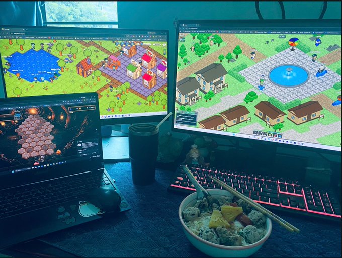 The image shows a desk with two computer monitors displaying a video game scene, an open laptop, and a bowl of food. The desktop monitor shows a map of a virtual world, while the laptop screen displays a game interface. A keyboard and mouse are also present on the desk. In front of the desk is a bowl filled with food, possibly snacks or a meal for someone to enjoy during breaks from gaming sessions.