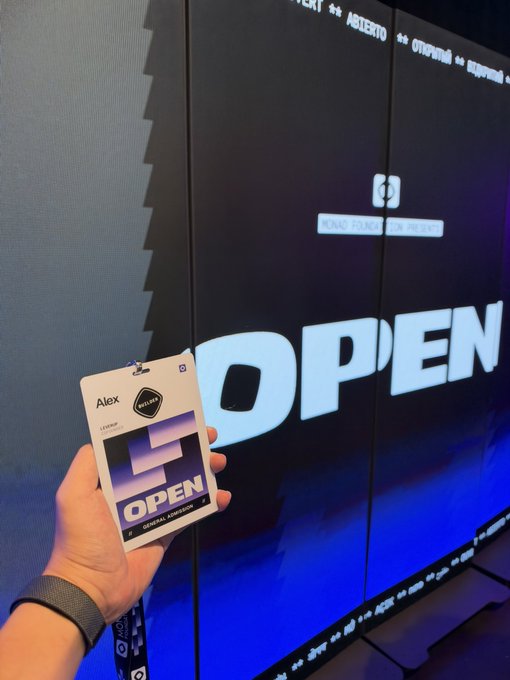 A hand holds an "Alex" credential tagged "OPEN" with a "Monad" logo, displayed in front of a large screen reading "OPEN," marking the start of the Monad blockchain event.