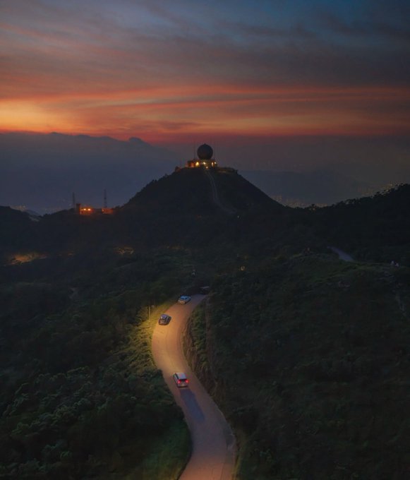 A scenic sunset view over a mountain with a radar dome on top and a winding road below where cars drive with their headlights on.