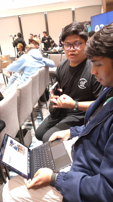 Two young men work on laptops in what appears to be a busy convention or workspace setting, with one wearing glasses and a black t-shirt while the other in a navy jacket focuses on his screen.