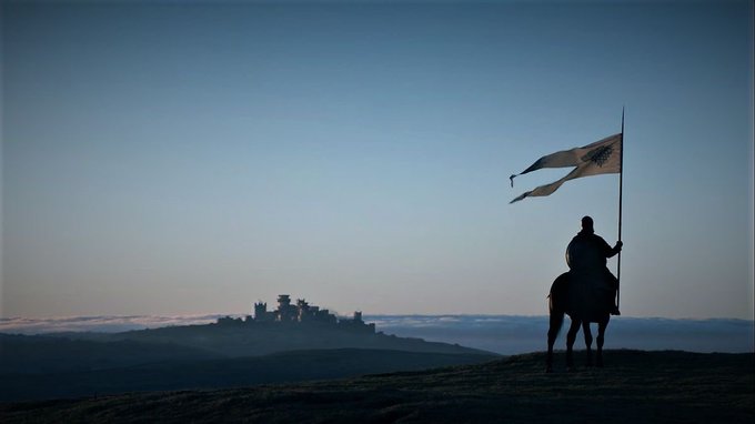 A silhouette of a knight on horseback holding a flag with a tree emblem stands on a hill, with a distant castle visible on the horizon at dusk.