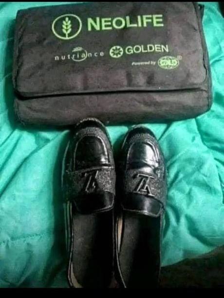 The image features a pair of black loafers with silver buckles, placed next to a black bag that has "NeoLife" written on it. The shoes are positioned on top of a bedspread or blanket, which suggests they might be in someone's bedroom. The main focus of the image is the black loafers and the black bag, both of which have the word "NeoLife" printed on them.