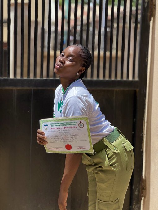 A young woman in a white t-shirt and olive-green pants proudly displays a "Certificate of Participation" from the Nigerian Dignity Drive Community Empowerment Service, posing playfully in front of a metal gate. The image reflects the post's theme of exhaustion and dedication, capturing her celebratory moment despite the late-night research efforts mentioned in the caption.