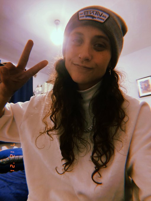 A selfie of a smiling young woman wearing a gray beanie and white sweatshirt, making a peace sign indoors, with a timestamp showing '1-21' in the corner.