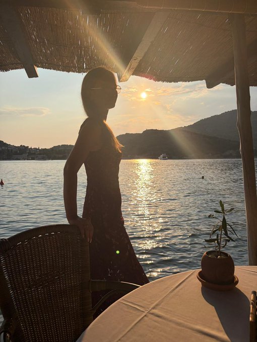 A woman in sunglasses stands silhouetted beneath a thatched canopy, gazing out at a golden sunset reflecting over calm waters with distant hills in the background. A small potted plant sits on the white tablecloth beside her, perfectly capturing the serene "Good evening" atmosphere of the seaside scene.