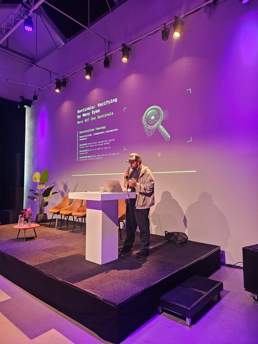 A man wearing a cap and glasses stands on a stage giving a presentation, with a large purple-lit screen behind him displaying "Sentinals: Verifying for Many Eyes Not WCT One Sentinels" alongside a magnifying glass graphic.